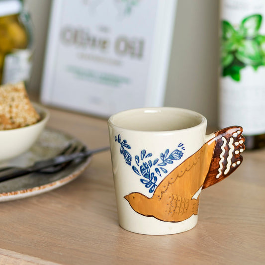 White mug with hand painted bird at a table setting. The bird wing is the handle