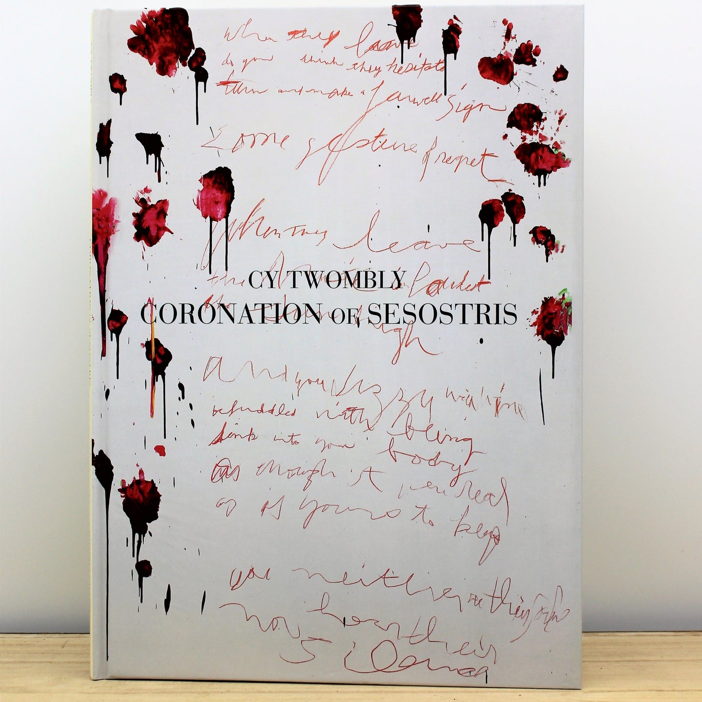 The image shows the cover of a book titled 'Cy Twombly: Coronation of Sesostris' with a abstract design resembling blood splatters over a white background, complementing the artistic content of the book which features photographs of Cy Twombly's artwork.