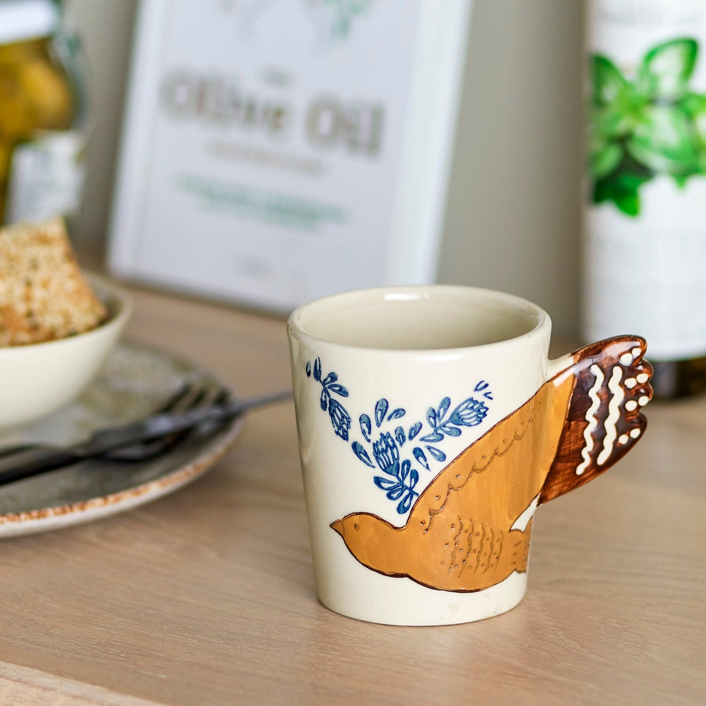 White mug with hand painted bird at a table setting. The bird wing is the handle 