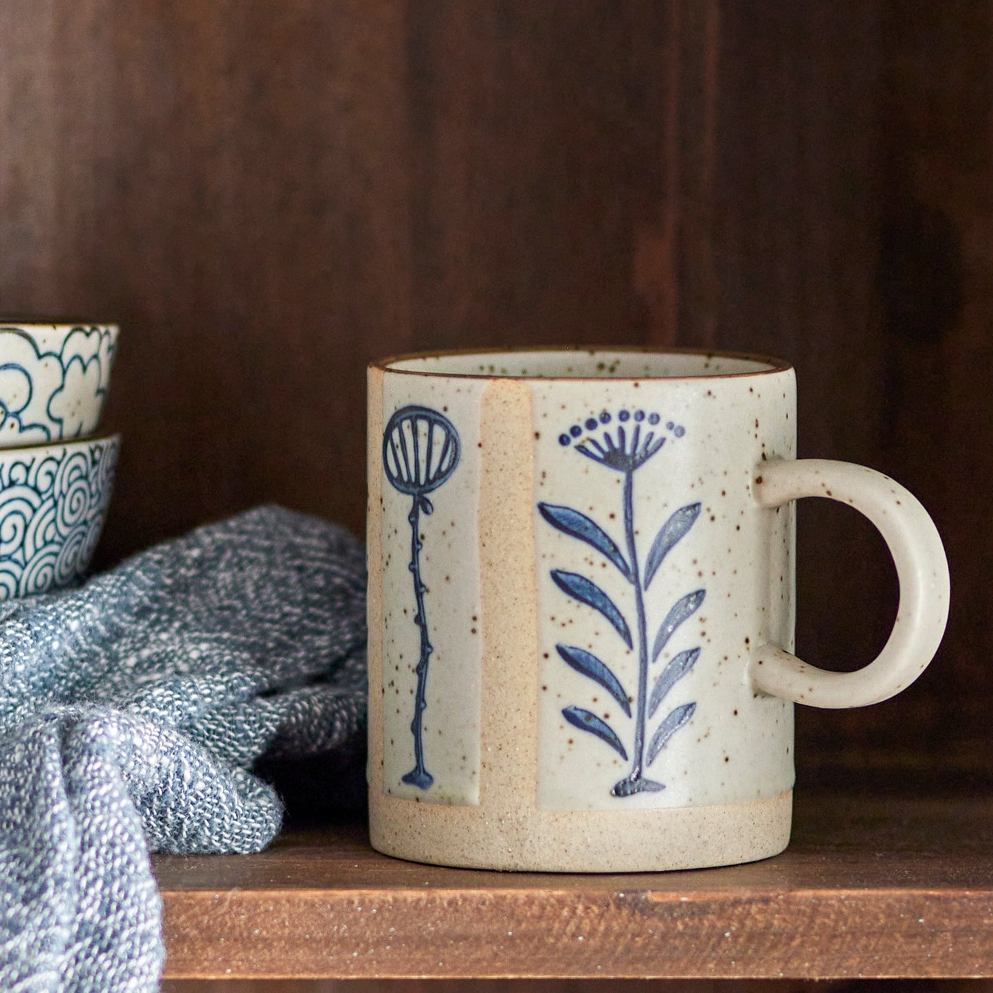 Small stoneware mug with blue floral detail placed on wooden shelf. 