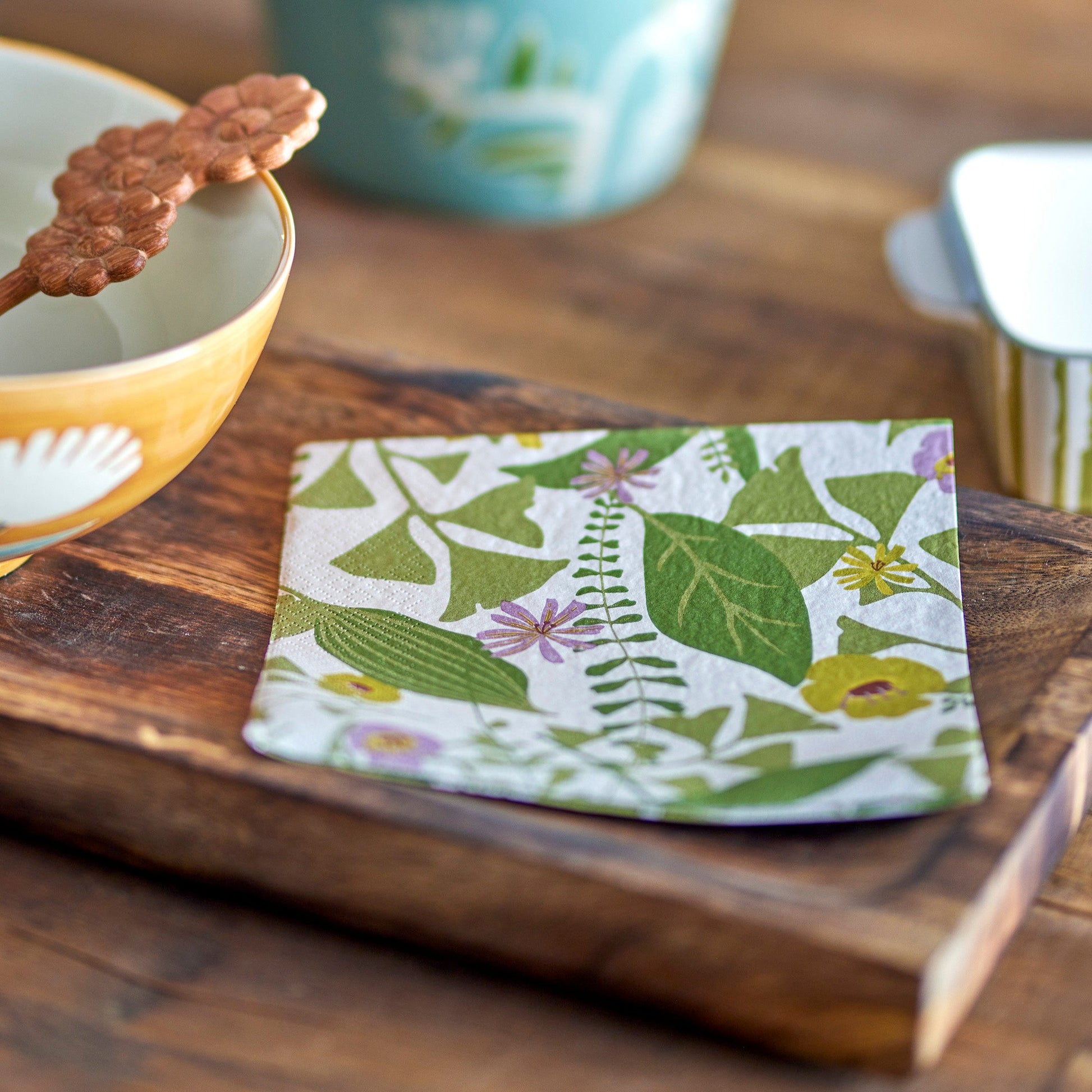 paper napkins with green foliage and pink and purple flowers on a wooden board surrounded by other tableware.