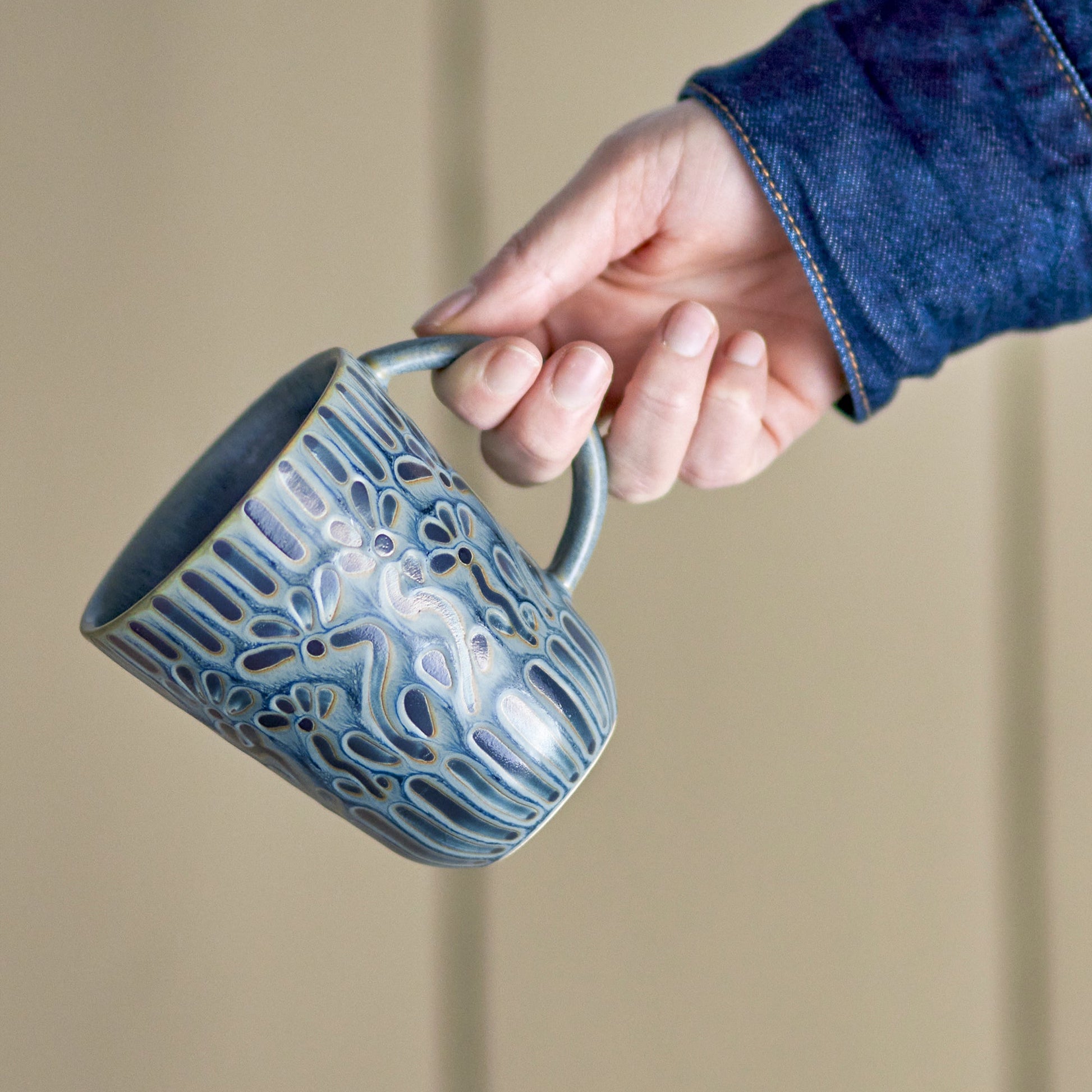 zoomed photo of person's hand holding mug. They are wearing a denim shirt. 