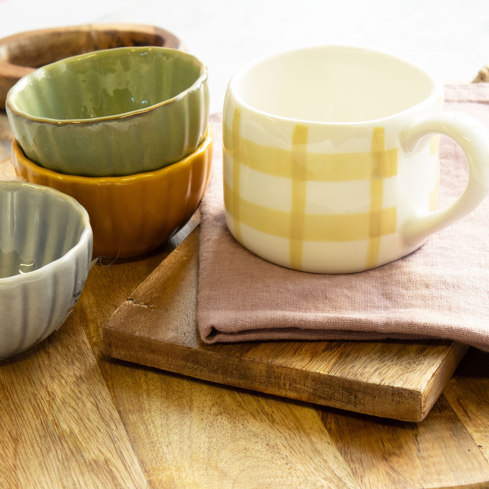 Yellow checked mug displayed on wood chopping boards. displayed next to a trio of decorative bowls. 